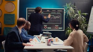 A diverse business team is engaged in a strategy meeting in a modern office space. A man in a dark suit is standing in front of a large digital screen displaying financial graphs and analytics, pointing at key data points. Three colleagues, seated around a white table, are attentively observing the presentation, holding documents and discussing insights. The workspace is decorated with plants, geometric wall panels, and a whiteboard filled with notes, creating a dynamic and collaborative atmosphere.