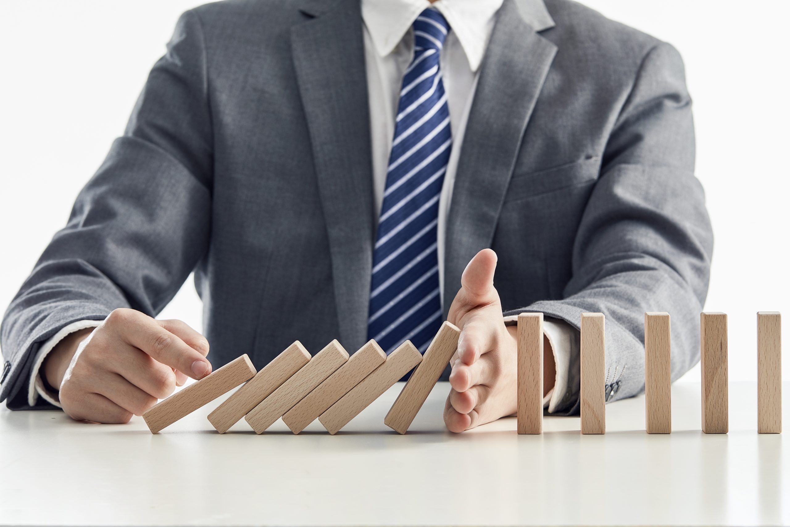 A businessman in a suit and striped tie is stopping a chain reaction of falling wooden domino blocks with his hand. The image symbolizes risk management, crisis prevention, and strategic intervention in business or finance. The background is plain white, emphasizing the professional setting and the concept of controlling unforeseen challenges.