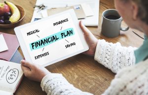 A person wearing a white knitted sweater is holding a tablet displaying a financial plan diagram with key aspects such as medical, insurance, expenses, and saving. The setting includes a wooden table with a cup of coffee, a notebook, and a bowl of fruit, suggesting a cozy and organized workspace. The image represents financial planning, budgeting, and investment management.