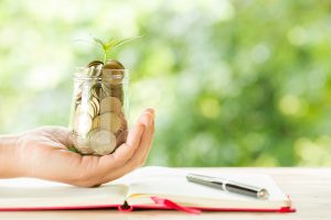 A hand is holding a glass jar filled with coins, from which a small green plant is growing, symbolizing financial growth and investment. The jar is placed above an open notebook with a pen resting on the pages, suggesting planning and saving for the future. The background is blurred with lush greenery, emphasizing the concept of sustainable financial growth and prosperity.