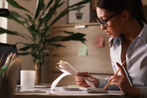 A woman wearing glasses is sitting at her desk in an office, carefully reviewing receipts while calculating her finances with a calculator, surrounded by office supplies and a cup of coffee.