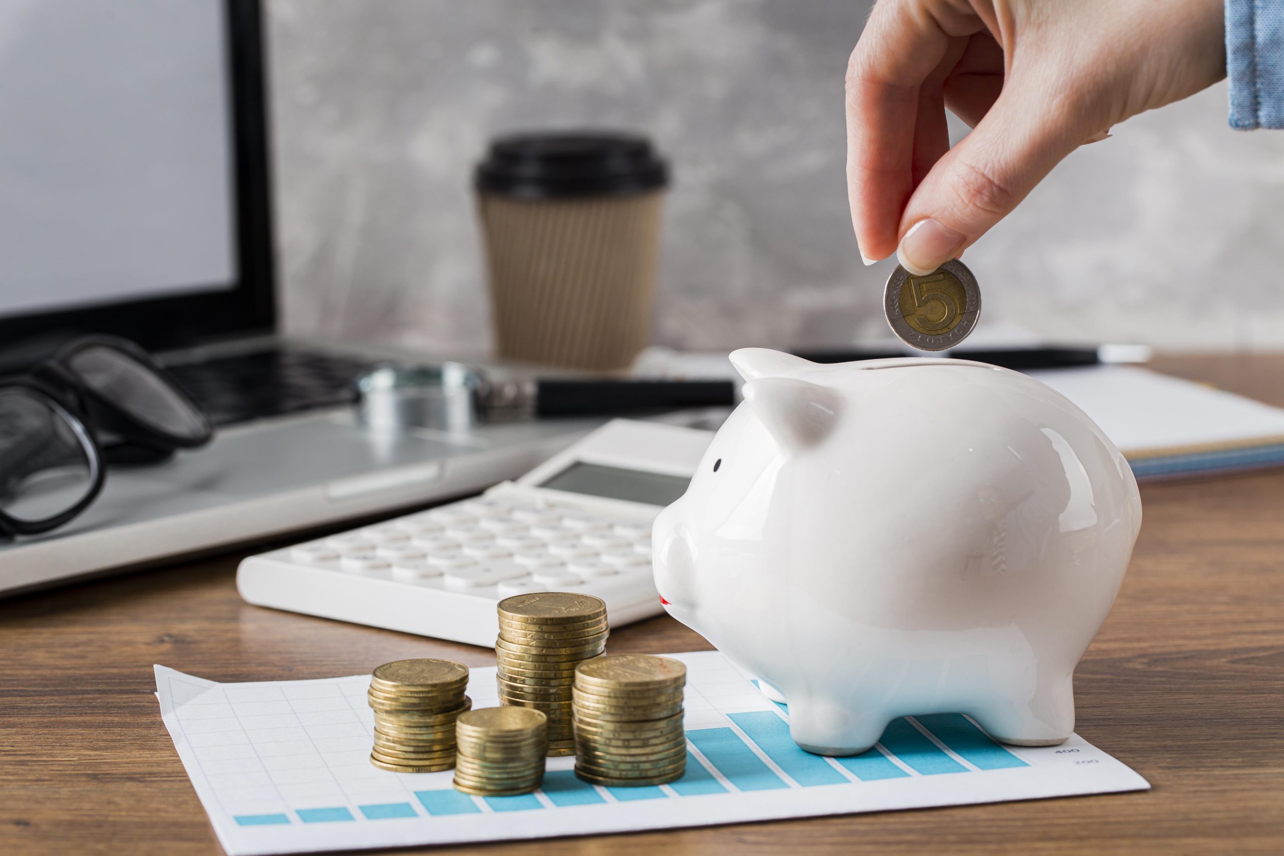 A person placing a coin into a piggy bank, surrounded by stacks of coins and a growth chart, symbolizing saving money and financial growth, with a laptop and calculator in the background.