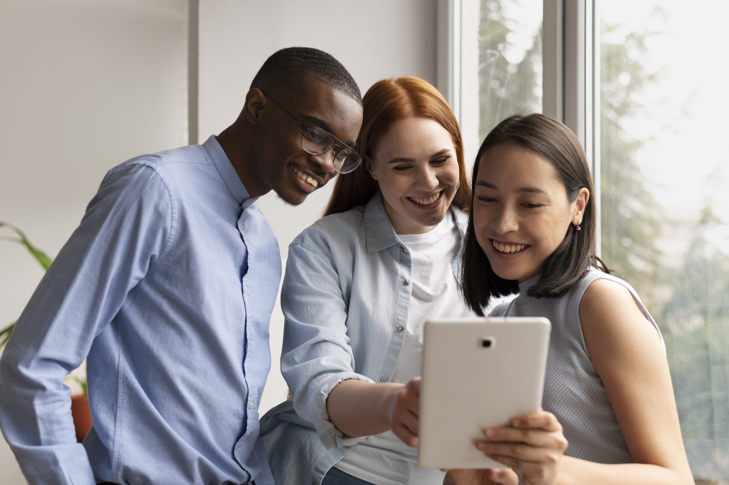 A group of three friends, a man and two women, smiling and looking at a tablet together in a bright, modern workspace, sharing a moment of social connection.