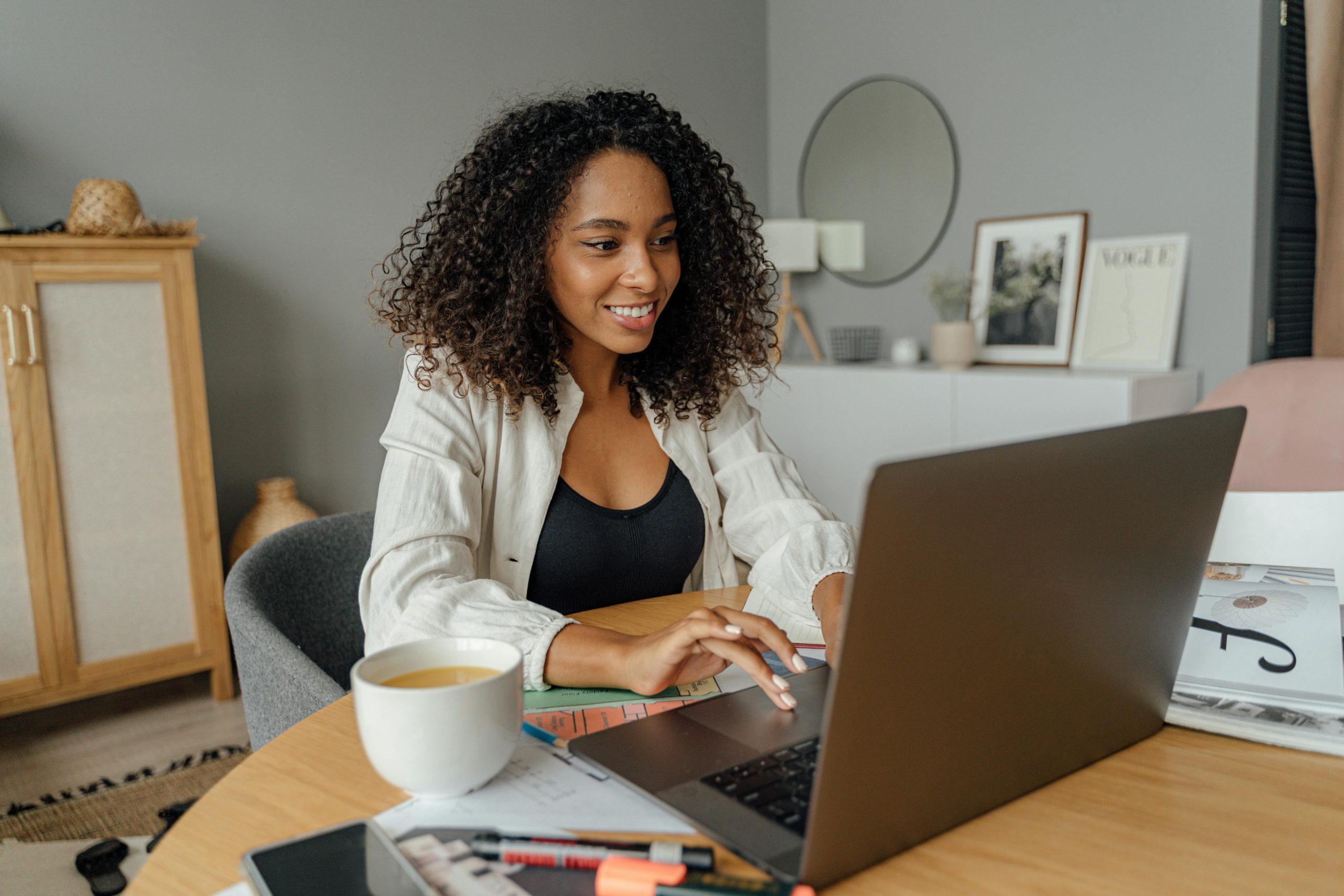 A woman with curly hair is smiling and working on a laptop at a desk in a cozy, modern workspace. She is surrounded by various work materials, including a cup of tea, a notebook, pens, and a phone.