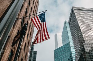 A close-up view of the United States flag flying from a flagpole in front of towering skyscrapers. The buildings have glass facades, and the sky is cloudy, providing a dramatic urban scene.