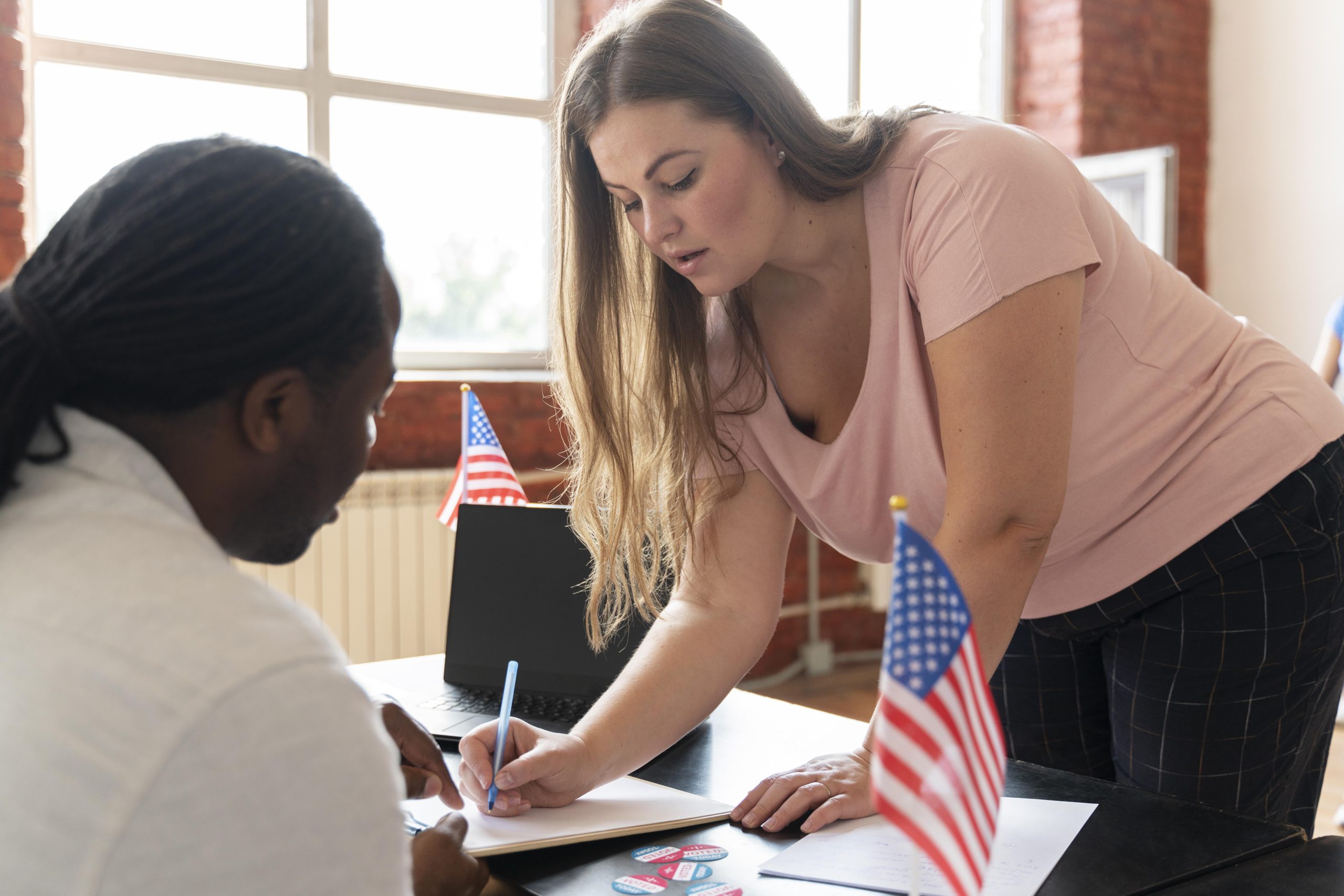 A woman with long hair is registering a vote while sitting at a table, with an American flag in the background, as a man watches her, in an office setting.