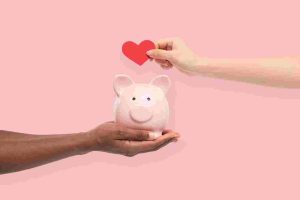 One hand holding a pink piggy bank while another places a red heart above it, symbolizing shared financial goals and savings strategies for couples.