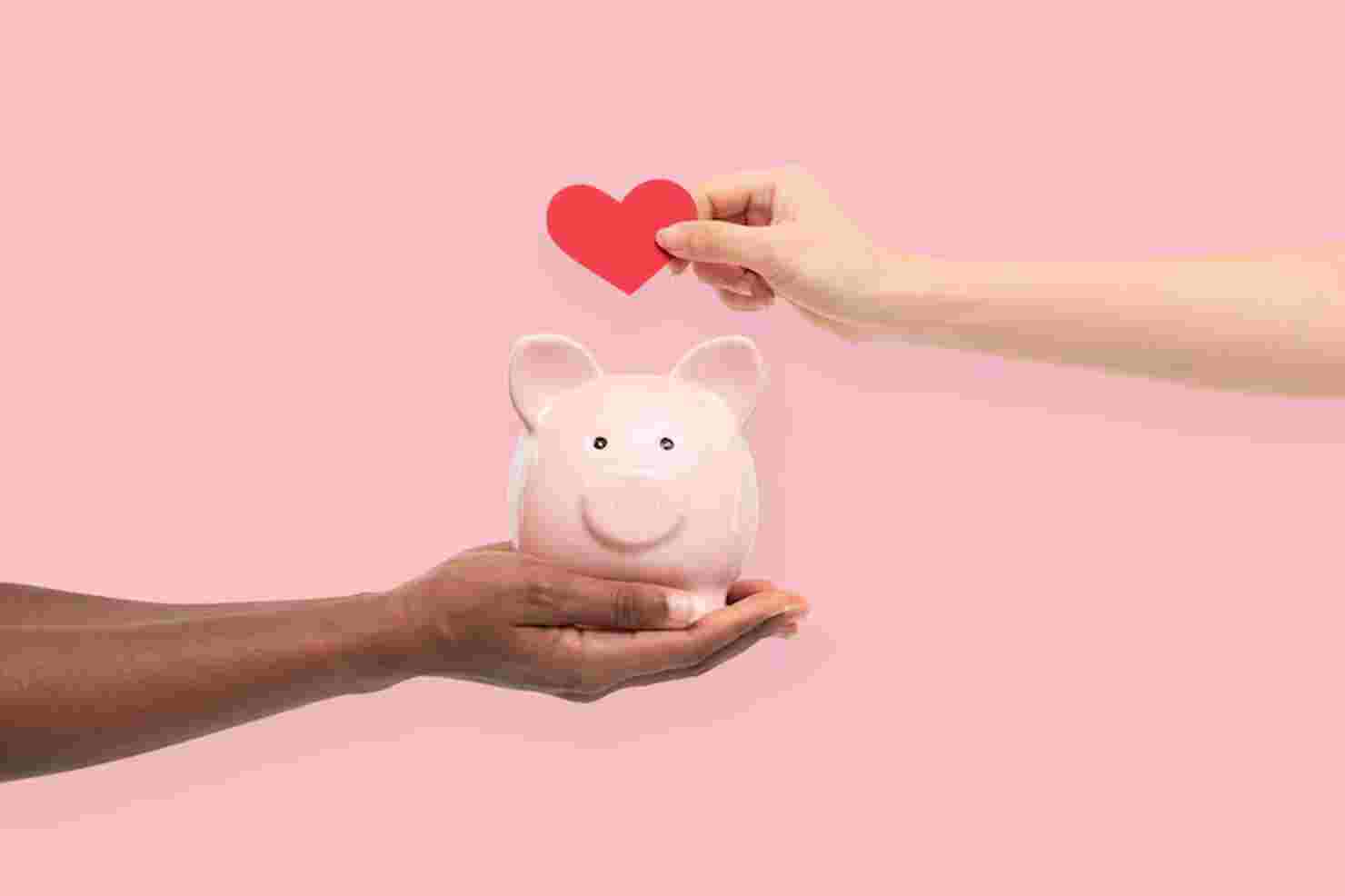 One hand holding a pink piggy bank while another places a red heart above it, symbolizing shared financial goals and savings strategies for couples.