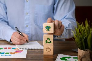 A person stacking wooden blocks with sustainability icons, including recycling and a green heart, while taking notes at a desk, symbolizing the principles and planning behind sustainable finance initiatives.