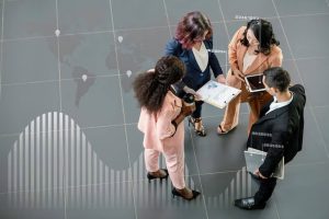 A diverse group of professionals gathered in a discussion over business documents and digital devices, with global data visuals overlaid on the floor, symbolizing the impact of globalization on the labor market and international workforce collaboration.