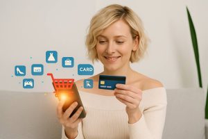A smiling blonde woman with short wavy hair using a smartphone and holding a credit card, surrounded by floating icons of online shopping, illustrating post-pandemic consumer trends such as increased digital purchases and mobile payment adoption.