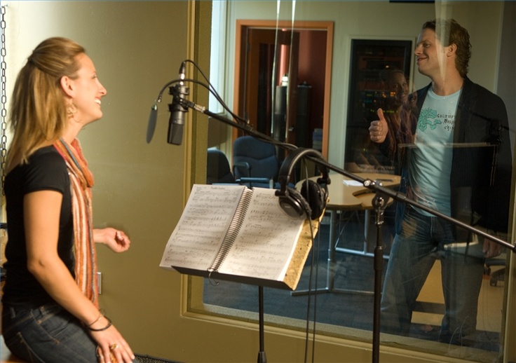 A smiling woman is inside a recording booth with sheet music, a microphone, and headphones, while a man stands outside the glass giving her a thumbs-up. The scene highlights collaboration in a music studio, reflecting the importance of investing in music and copyright to protect creative work.