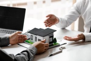 Two business professionals discussing a miniature house model on a desk with documents and keys, symbolizing investment opportunities and management of short-term rental properties.
