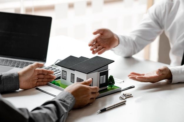 Two business professionals discussing a miniature house model on a desk with documents and keys, symbolizing investment opportunities and management of short-term rental properties.