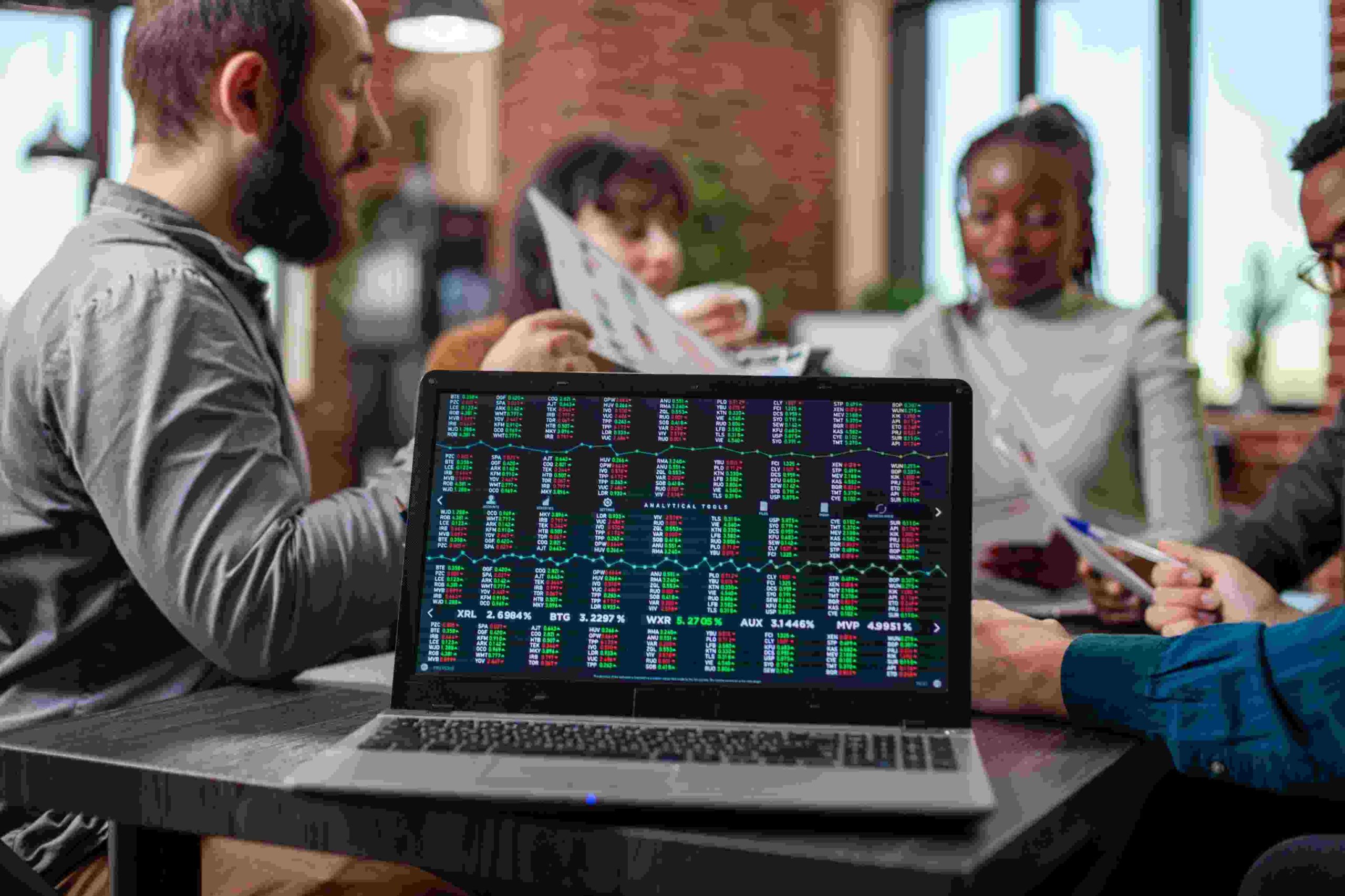 A group of people sitting around a table analyzing charts and financial reports, with a laptop screen showing stock market data in the foreground, symbolizing the collaborative strategies of investment clubs.